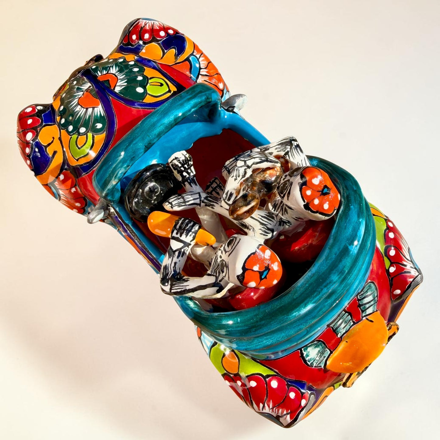 A colorful hand-painted ceramic Mexican-style convertible car figurine with Day of the Dead skeleton characters inside, shown on a neutral beige background. Top view of the car showing passengers and steering wheel.