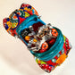 A colorful hand-painted ceramic Mexican-style convertible car figurine with Day of the Dead skeleton characters inside, shown on a neutral beige background. Top view of the car showing passengers and steering wheel.