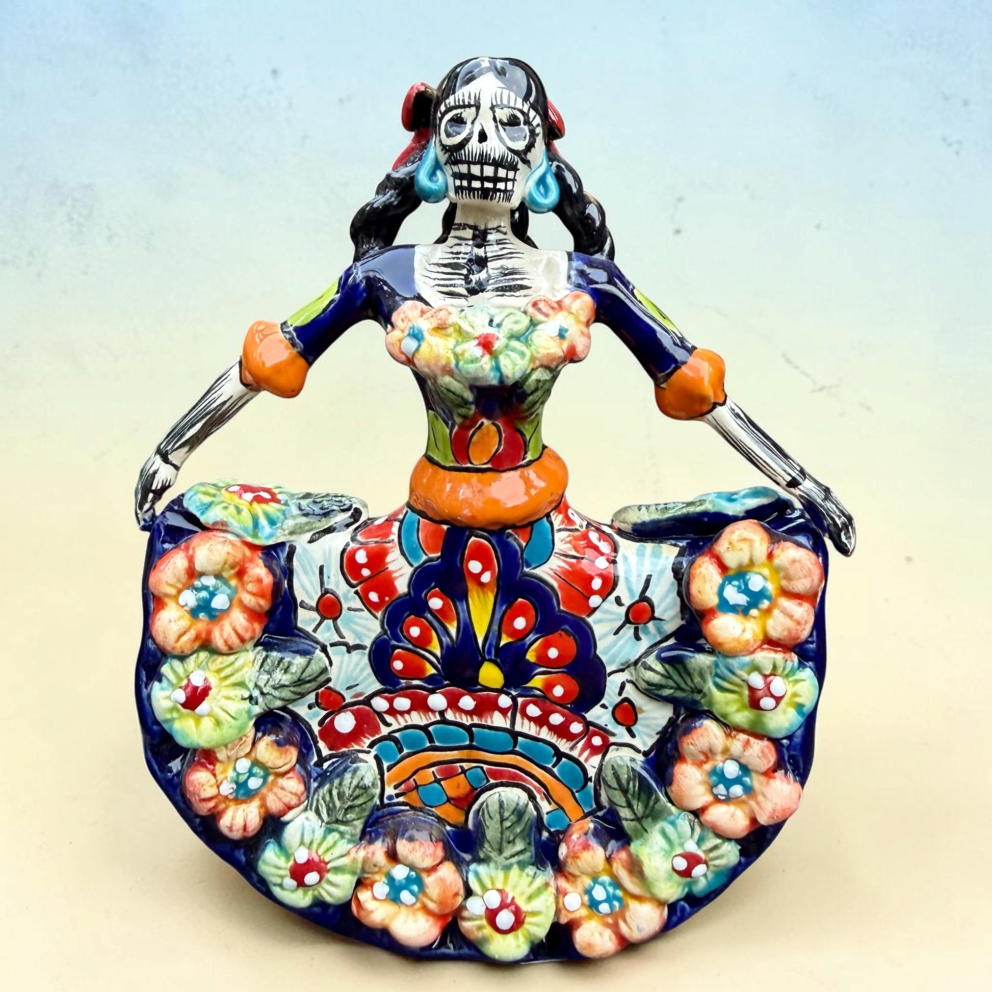 A colorful talavera Day of the Dead (Día de los Muertos) dancing skeleton ballerina figurine. The figurine features black hair in braids wearing a vibrant multicolor dress with floral patterns and orange accents, set against a light blue and beige gradient background.