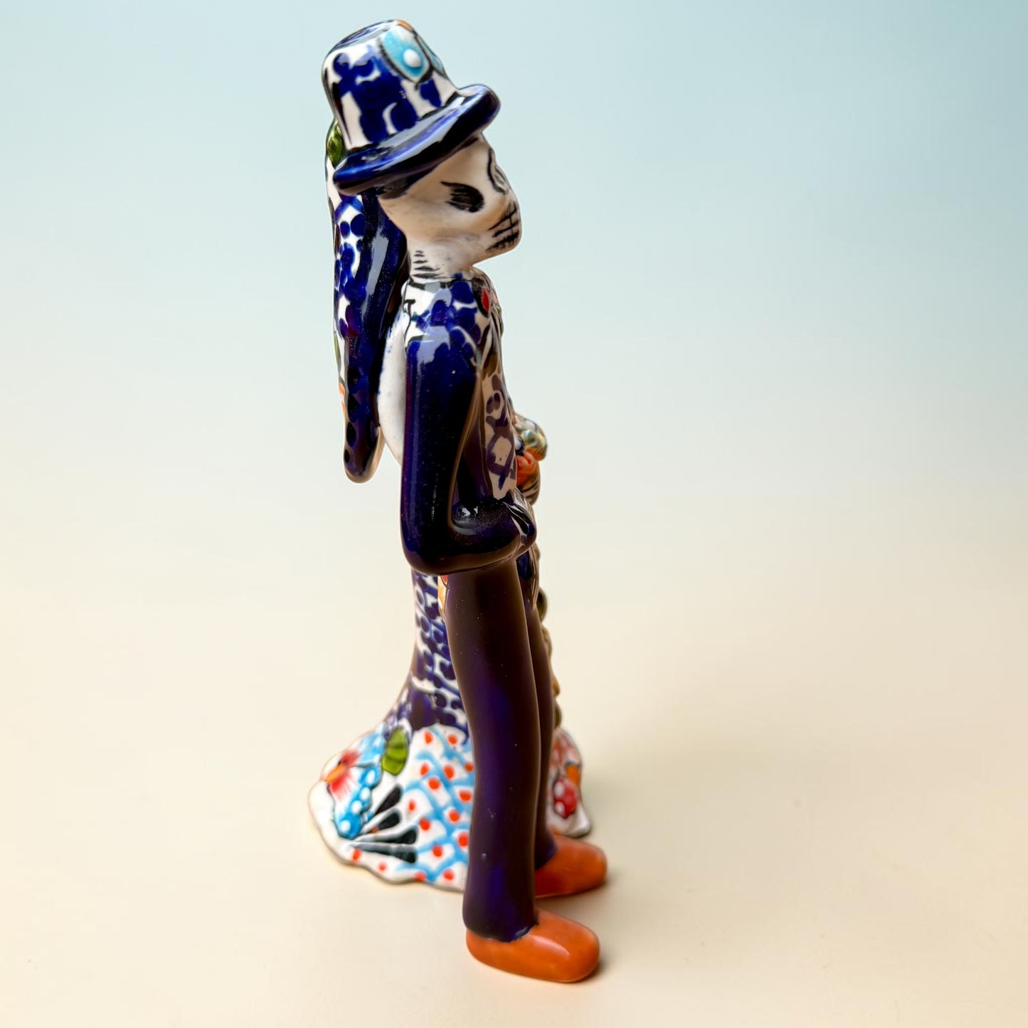 A hand-painted Talavera Day of the Dead bride and groom figurine. The groom is dressed in a blue suit with white patterns, a black bow tie, and an orange-red bow tie accent, wearing a blue hat. Left side view featuring groom.