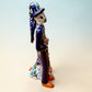 A hand-painted Talavera Day of the Dead bride and groom figurine. The groom is dressed in a blue suit with white patterns, a black bow tie, and an orange-red bow tie accent, wearing a blue hat. Left side view featuring groom.