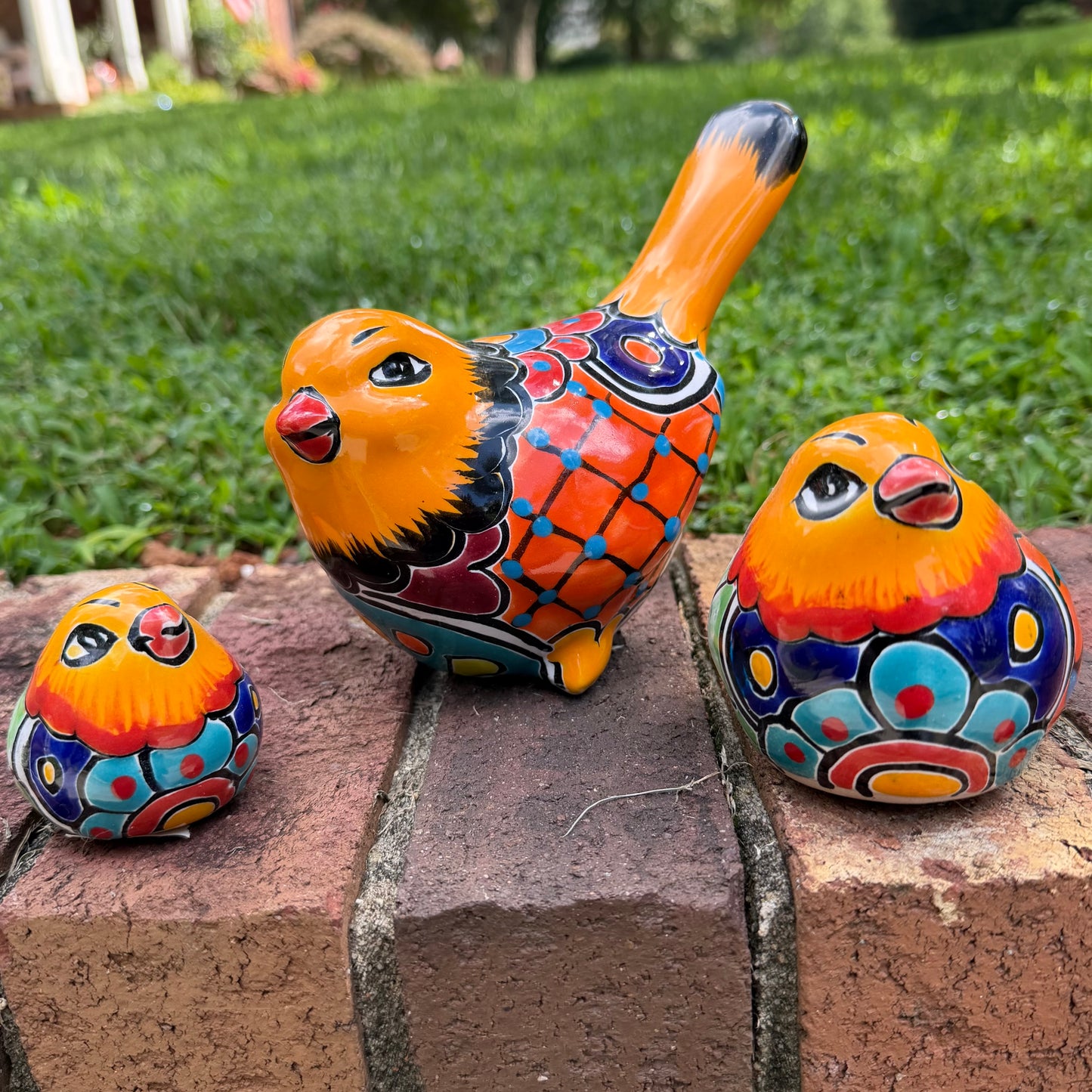 Three hand-painted ceramic bird figurines in yellow, orange  blue, red, and black, displayed on a brick surface with a green grass background.