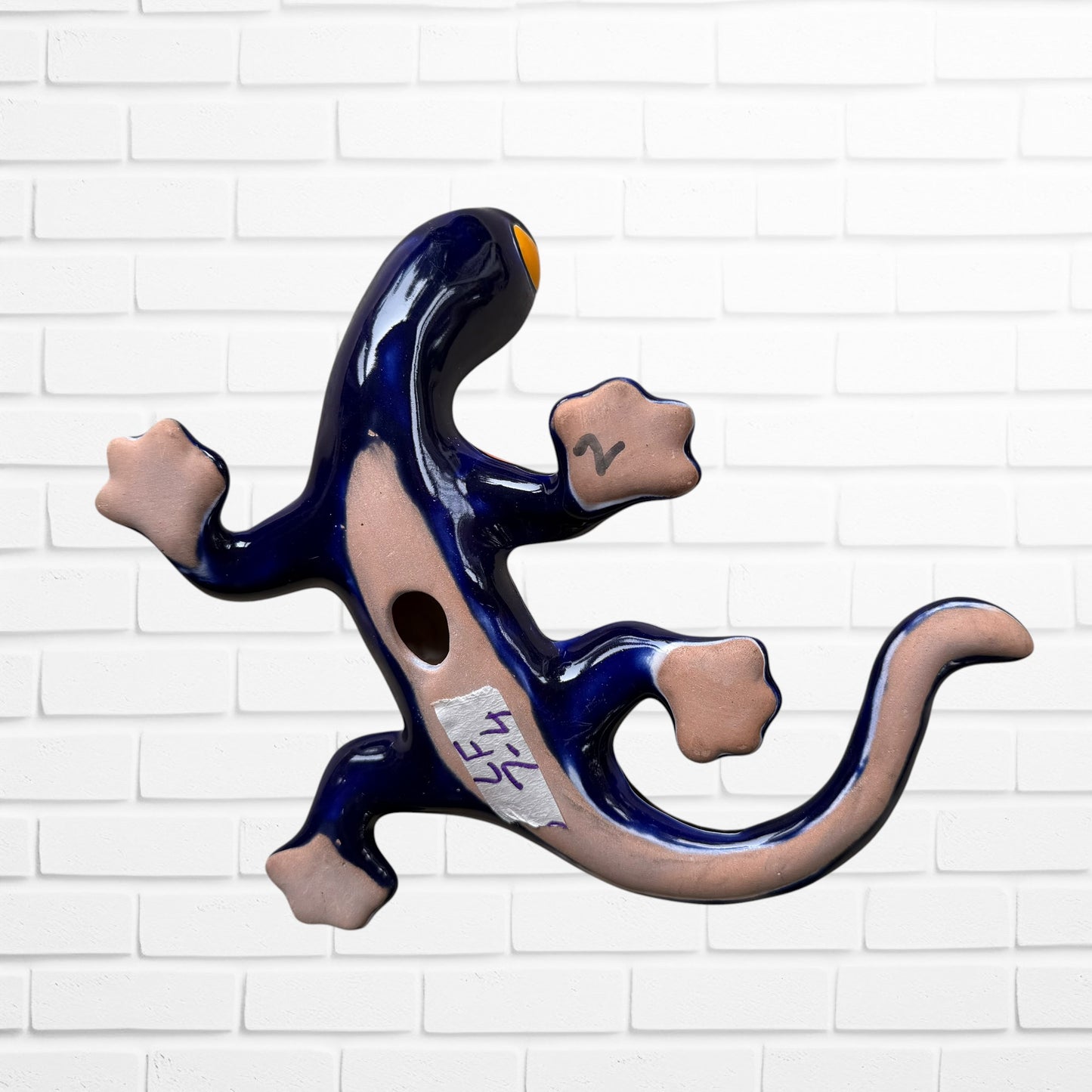 A colorful, glossy lizard wall sculpture with 
a vibrant mosaic pattern of blue, orange, yellow, green, red, and black, mounted on a white brick wall bottom view with hanging hole