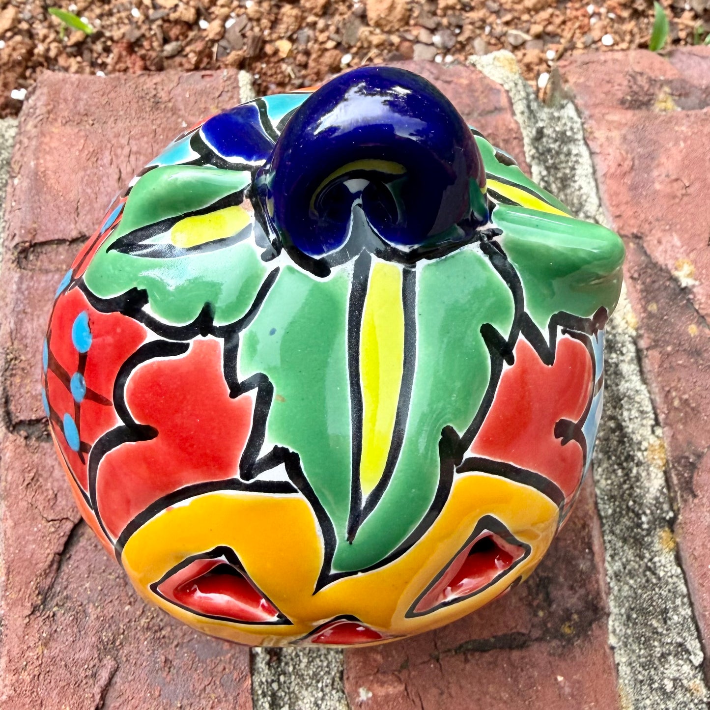 One hand-painted Talavera ceramic jack-o'-lantern pumpkins in bright orange, yellow, red, green, and blue, sitting on a brick ledge with a blurred green lawn and house in the background, top stem view.