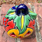 One hand-painted Talavera ceramic jack-o'-lantern pumpkins in bright orange, yellow, red, green, and blue, sitting on a brick ledge with a blurred green lawn and house in the background, top stem view.