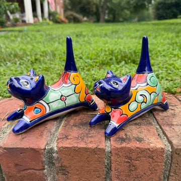 Two cobalt blue with multi-color designs hand-painted talavera ceramic cat figurines resting on a brick ledge with a green grassy lawn right side view.