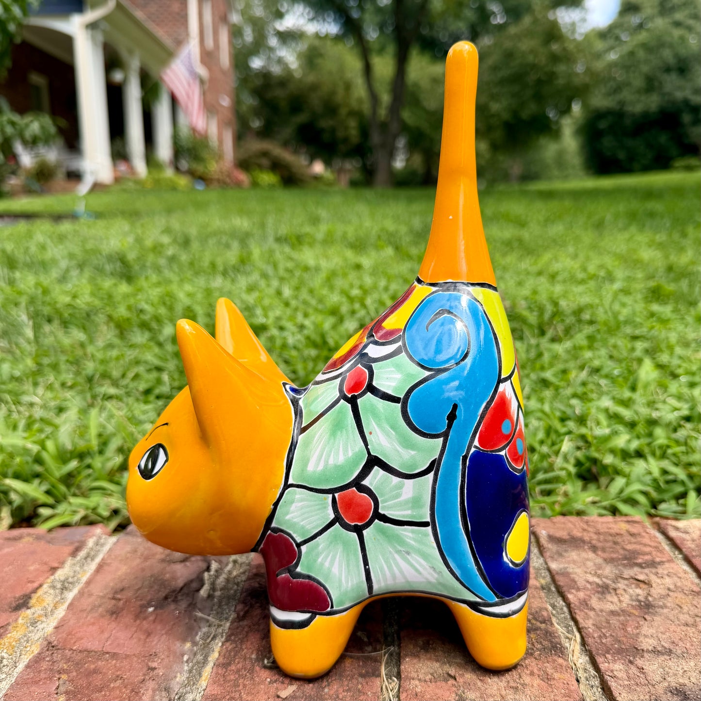 A hand-painted ceramic cat figurine in bright yellow with red, blue, and beige patterns, standing on a brick surface with a blurred green lawn and house in the background, right side view of cat.