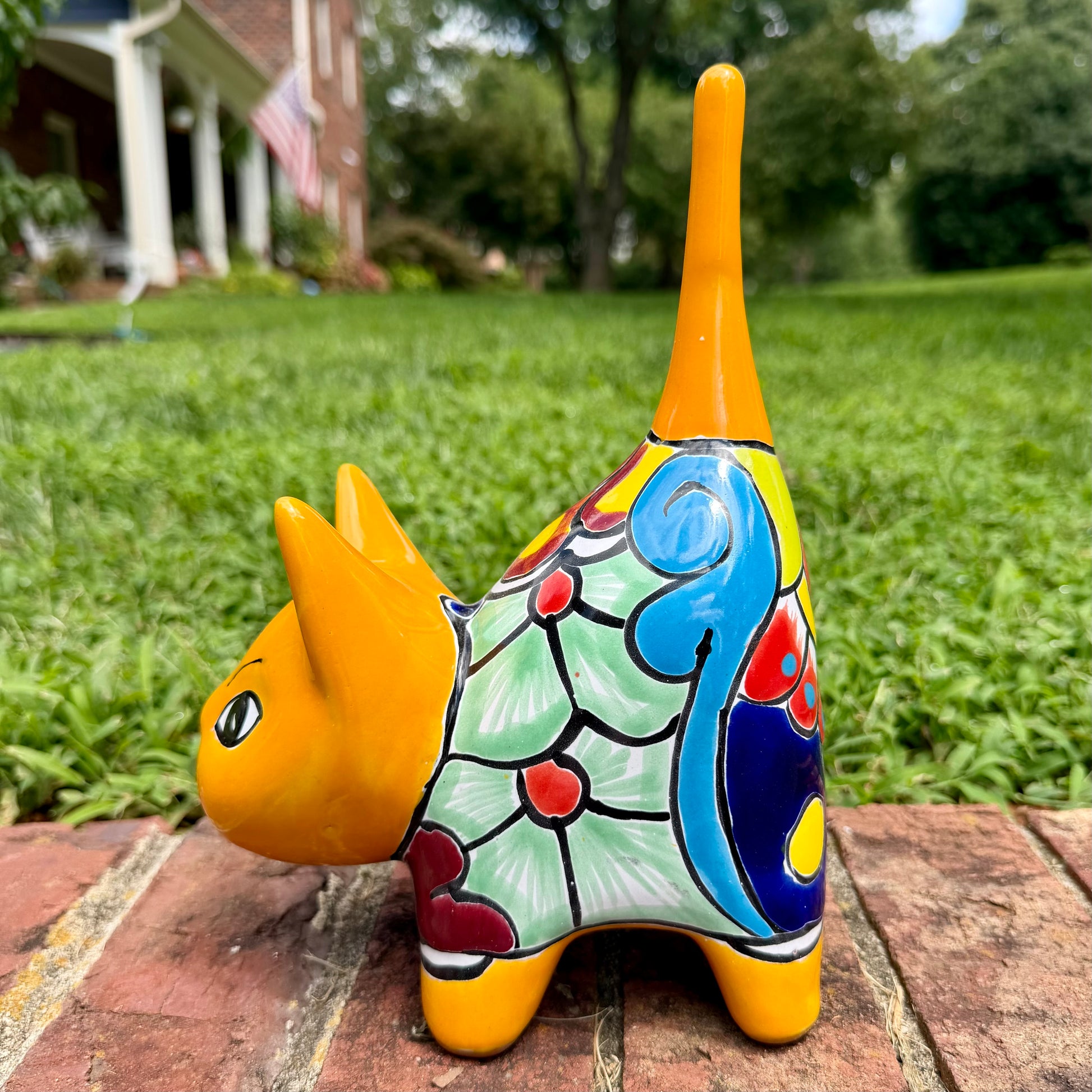 A hand-painted ceramic cat figurine in bright yellow with red, blue, and beige patterns, standing on a brick surface with a blurred green lawn and house in the background, right side view of cat.