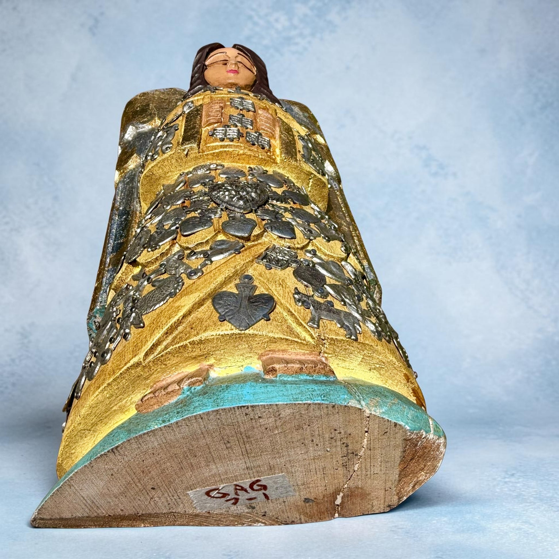 Milagros wooden guardian angel figurine wearing a yellow robe with gold and silver metallic details, standing on a light blue textured background, bottom?