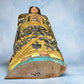 Milagros wooden guardian angel figurine wearing a yellow robe with gold and silver metallic details, standing on a light blue textured background, bottom?