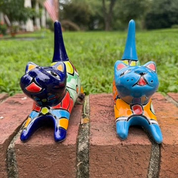 Two colorful hand-painted ceramic cats sitting on a brick ledge with a green lawn and trees in the background. One is cobalt  blue and the other is turquoise, both have traditional multi-color  talavera designs. his is the front face view.