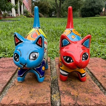 Two hand-painted Mexican-style ceramic cat figurines, one blue and one red, decorated with colorful geometric patterns, standing on a brick surface outdoors front view