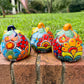 Three hand-painted Talavera ceramic jack-o'-lantern pumpkins with orange faces and yellow, blue, and red green designs, sitting on a brick path in a grassy backyard, back view.