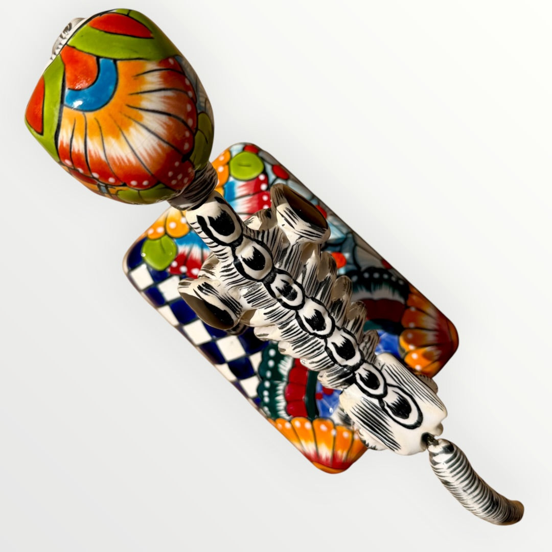 A talavera catrina dog skeleton figurine with colorful hand-painted floral and geometric patterns, standing on a matching decorated base against a plain white background. Top view of catrina dog.