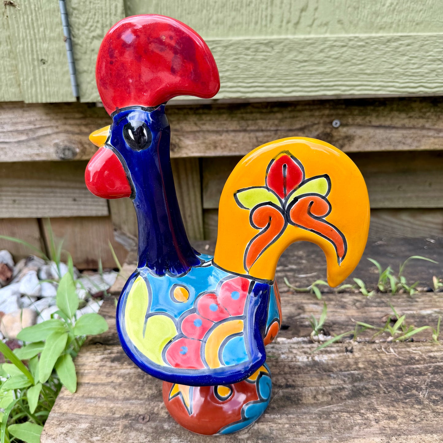 A colorful ceramic rooster figurine standing on a weathered wooden surface outdoors, with a green wooden wall behind it. The rooster is decorated with bright red, yellow, blue, and orange painted details, right side view.