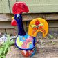 A colorful ceramic rooster figurine standing on a weathered wooden surface outdoors, with a green wooden wall behind it. The rooster is decorated with bright red, yellow, blue, and orange painted details, right side view.