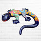A colorful Mexican Talavera wall sculpture of a lizard, featuring a vibrant multicolor pattern with dark blue, orange, red, green, and yellow, mounted on a white brick wall, left side view.