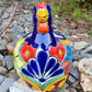 A colorful ceramic chicken-shaped planter with a glossy hand-painted finish, placed outdoors on a bed of small gray rocks and greenery, front face view.