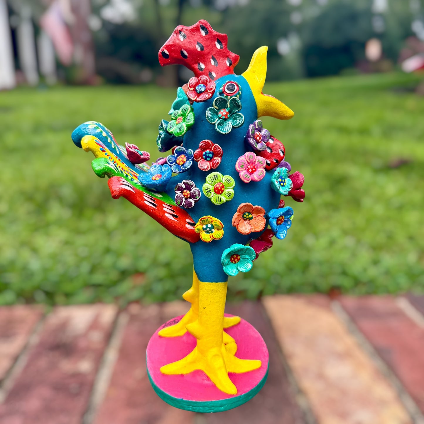 A colorful decorative rooster folk art sculpture with a blue body, yellow legs and beak, and many bright flower ornaments, standing on a pink circular base outdoors on a brick path with a blurred green lawn behind it. Left side rooster view. 