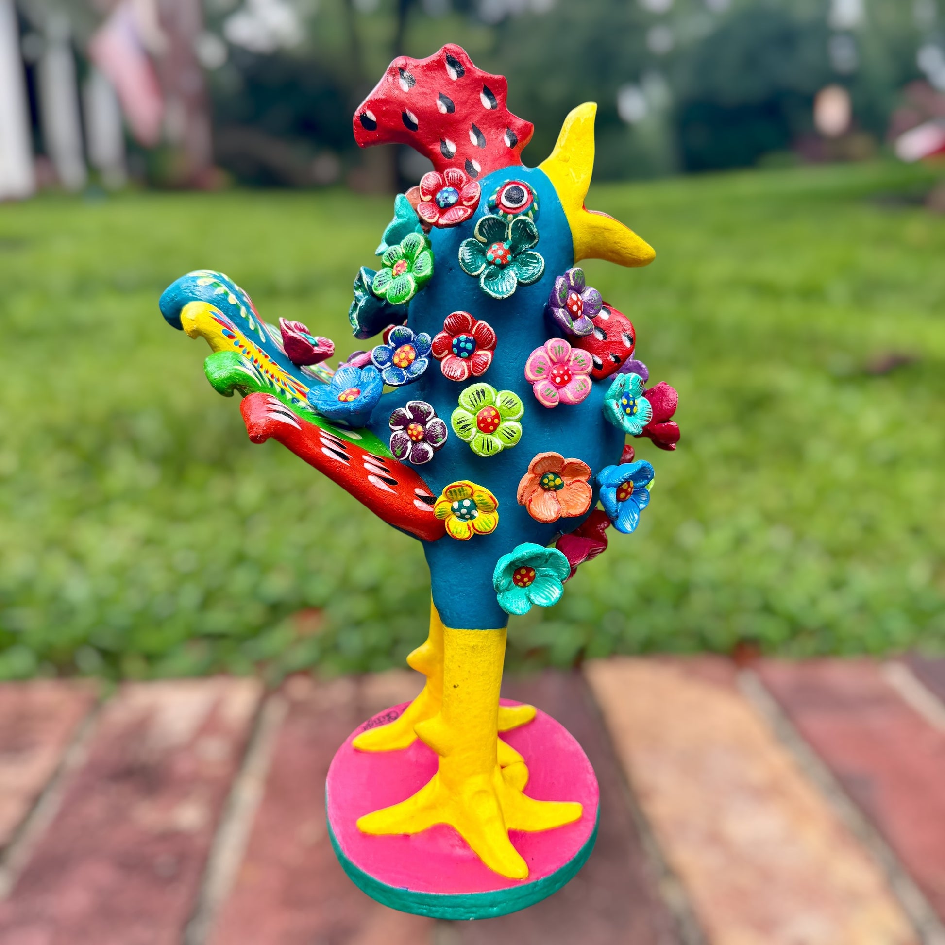 A colorful decorative rooster folk art sculpture with a blue body, yellow legs and beak, and many bright flower ornaments, standing on a pink circular base outdoors on a brick path with a blurred green lawn behind it. Left side rooster view. 