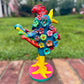 A colorful decorative rooster folk art sculpture with a blue body, yellow legs and beak, and many bright flower ornaments, standing on a pink circular base outdoors on a brick path with a blurred green lawn behind it. Left side rooster view. 