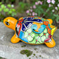 A glossy ceramic turtle figurine with a bright yellow body and a highly decorated shell in red, blue, yellow, black, and white, resting on a stone surface with a blurred garden background, right side view.