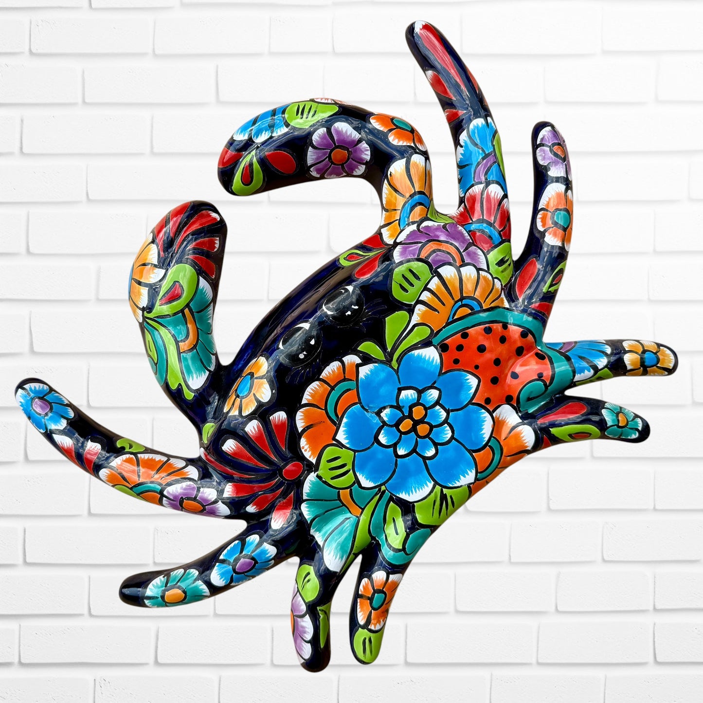 A colorful talavera crab wall art sculpture decorated with vibrant floral patterns, shown against a white brick wall background.