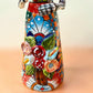 A Talavera Day of the Dead (Día de los Muertos) Frida figurine  with a monkey on her shoulder and a red parrot on her other shoulder, set against a light blue gradient background. Close-up of floral dress detail.