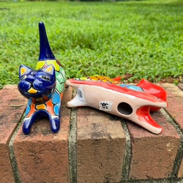 Two colorful ceramic cat figurines, one blue and one red, decorated with geometric patterns, displayed on a brick ledge with a green lawn in the background view showing bottom of the figurine.