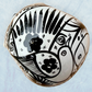 A decorative skull sculpture with intricate black sugar skull style patterns painted on an off-white base, set against a light blue textured background, top view.