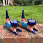 Two cobalt blue with multi-color designs hand-painted talavera ceramic cat figurines resting on a brick ledge with a green grassy lawn in the background.