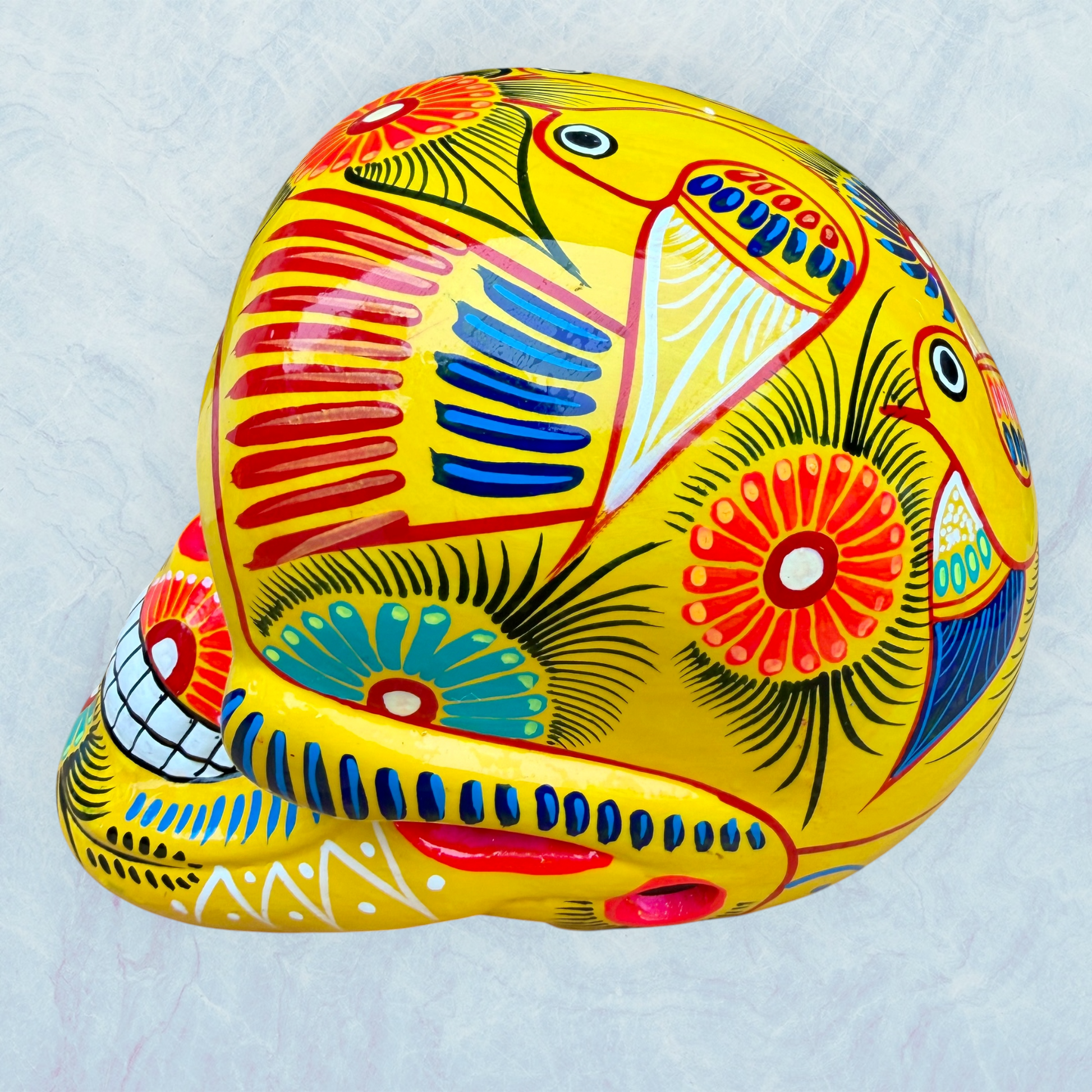A vibrant yellow sugar skull decorated with colorful floral and geometric patterns in blue, red, black, and white, set against a light blue textured background, right side view.