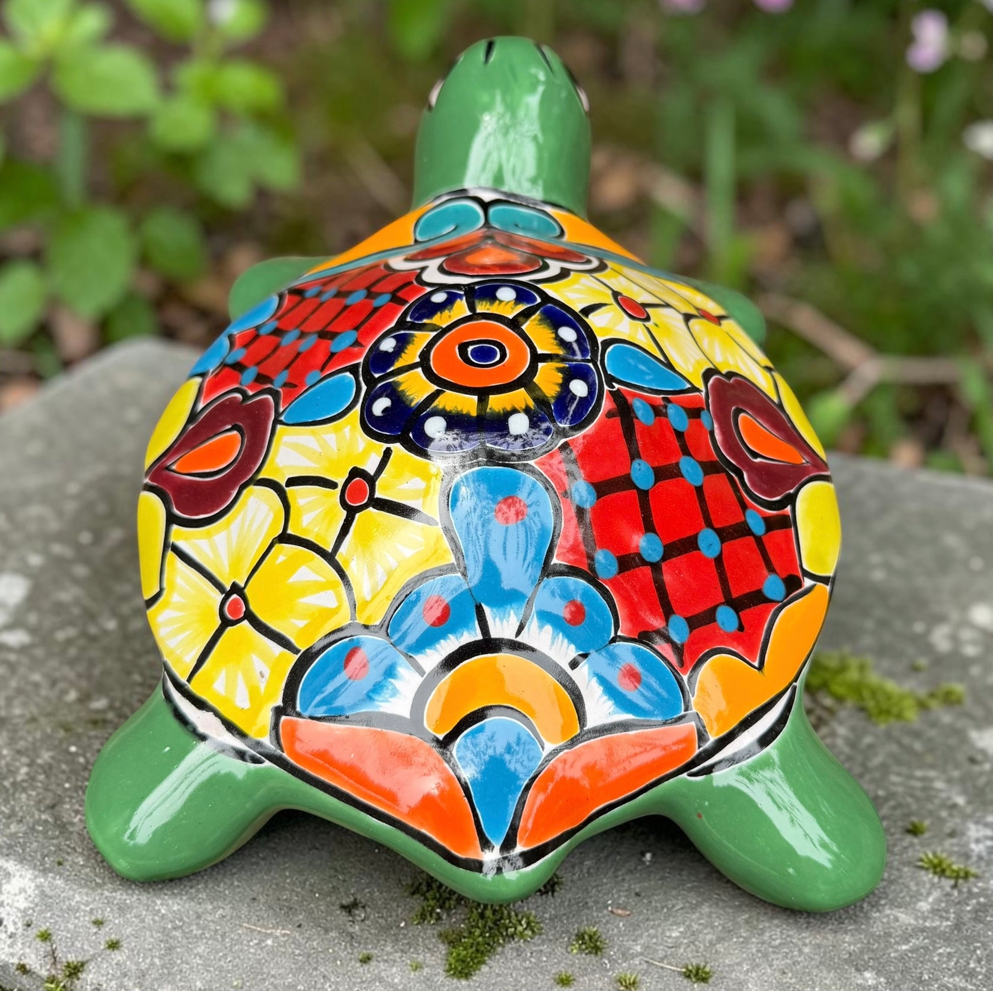 A hand-painted ceramic turtle figurine resting on a gray stone surface with a blurred green garden background. The turtle features a bright green head and legs, and a highly detailed, multicolored shell with yellow, red, orange, blue, and black patterns, back view of Shell.