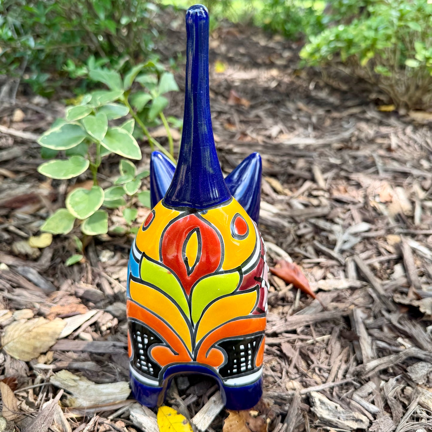 A colorful ceramic cat figurine with a glossy blue head and tail, yellow ears and paws, and a patterned body, standing outdoors on mulch with green plants in the background, back view of cat.