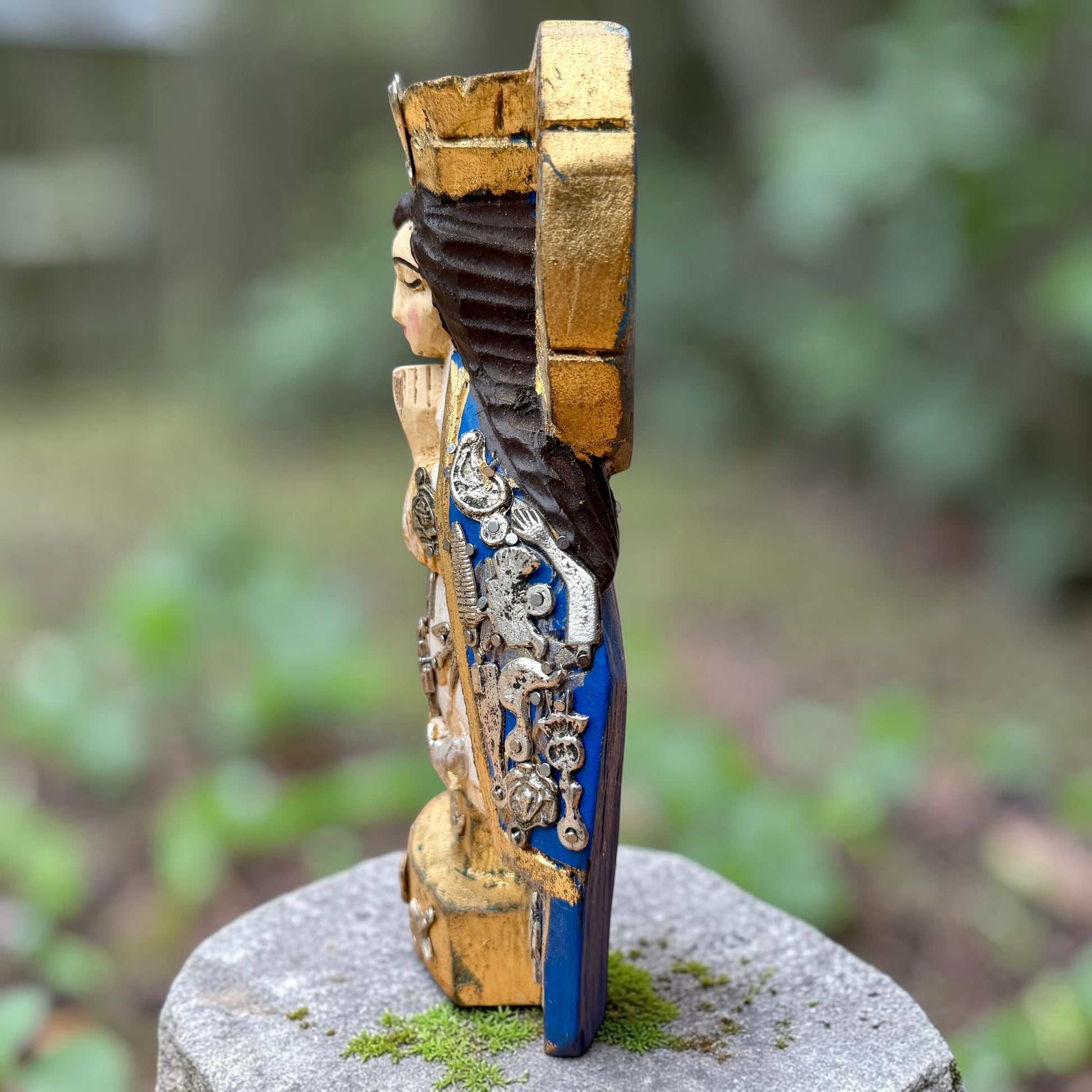 A Milagros figurine of Virgin Mary Salud, decorated with gold leaf, dark brown hair, and a vibrant blue patterned garment, displayed on a stone pedestal with a blurred garden background. This is the right side view.