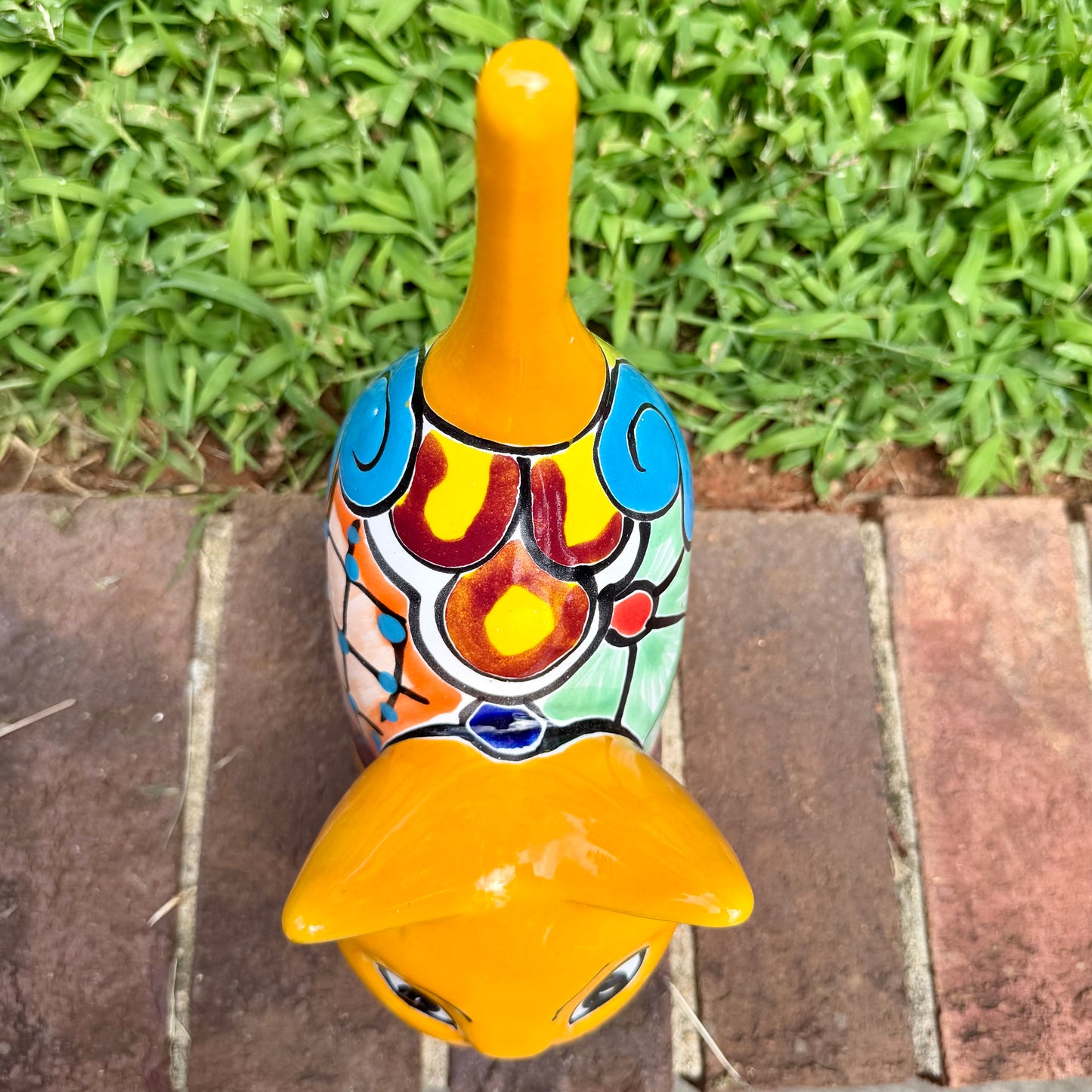 A hand-painted ceramic cat figurine in bright yellow with red, blue, and beige patterns, standing on a brick surface with a blurred green lawn and house in the background, top view of cat.