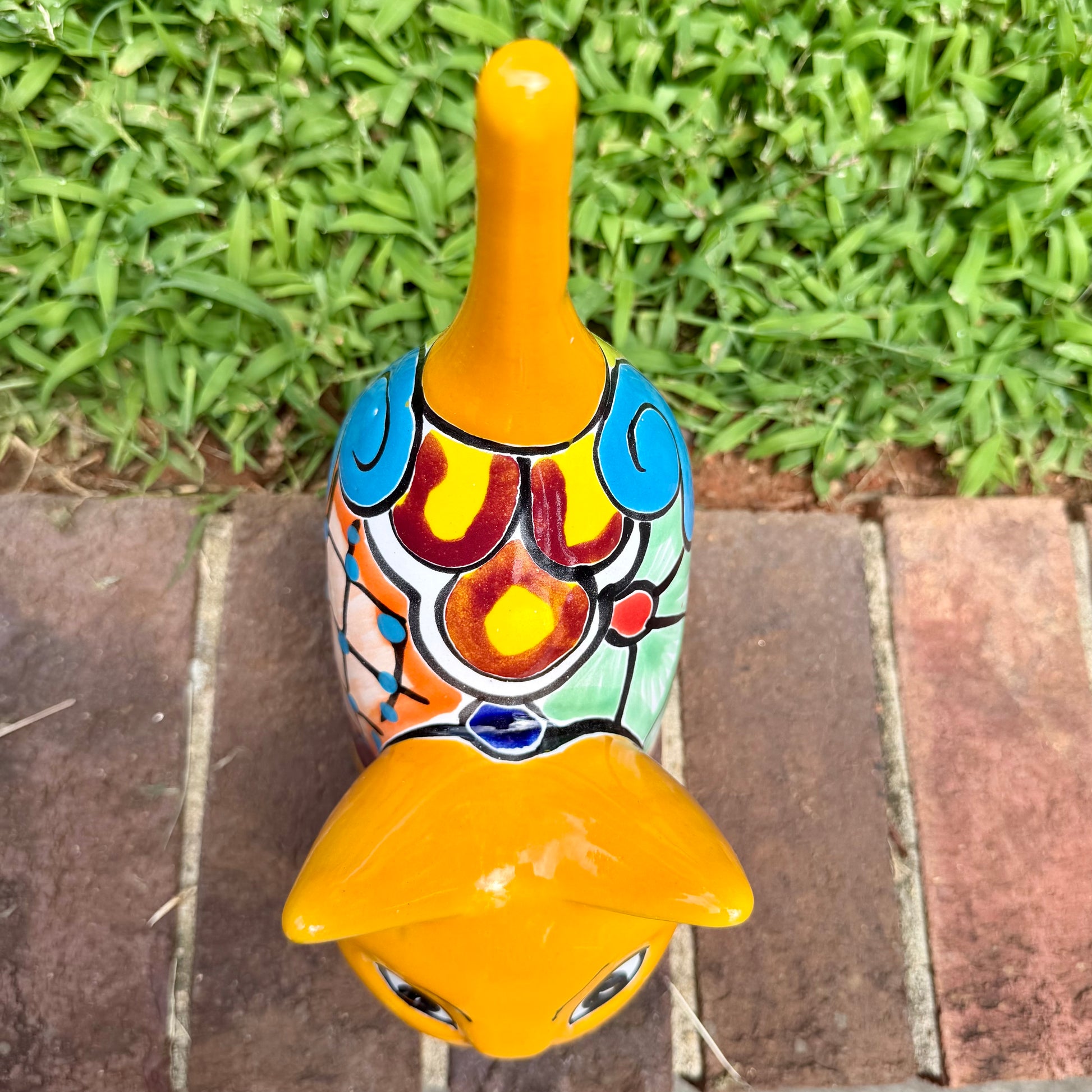 A hand-painted ceramic cat figurine in bright yellow with red, blue, and beige patterns, standing on a brick surface with a blurred green lawn and house in the background, top view of cat.