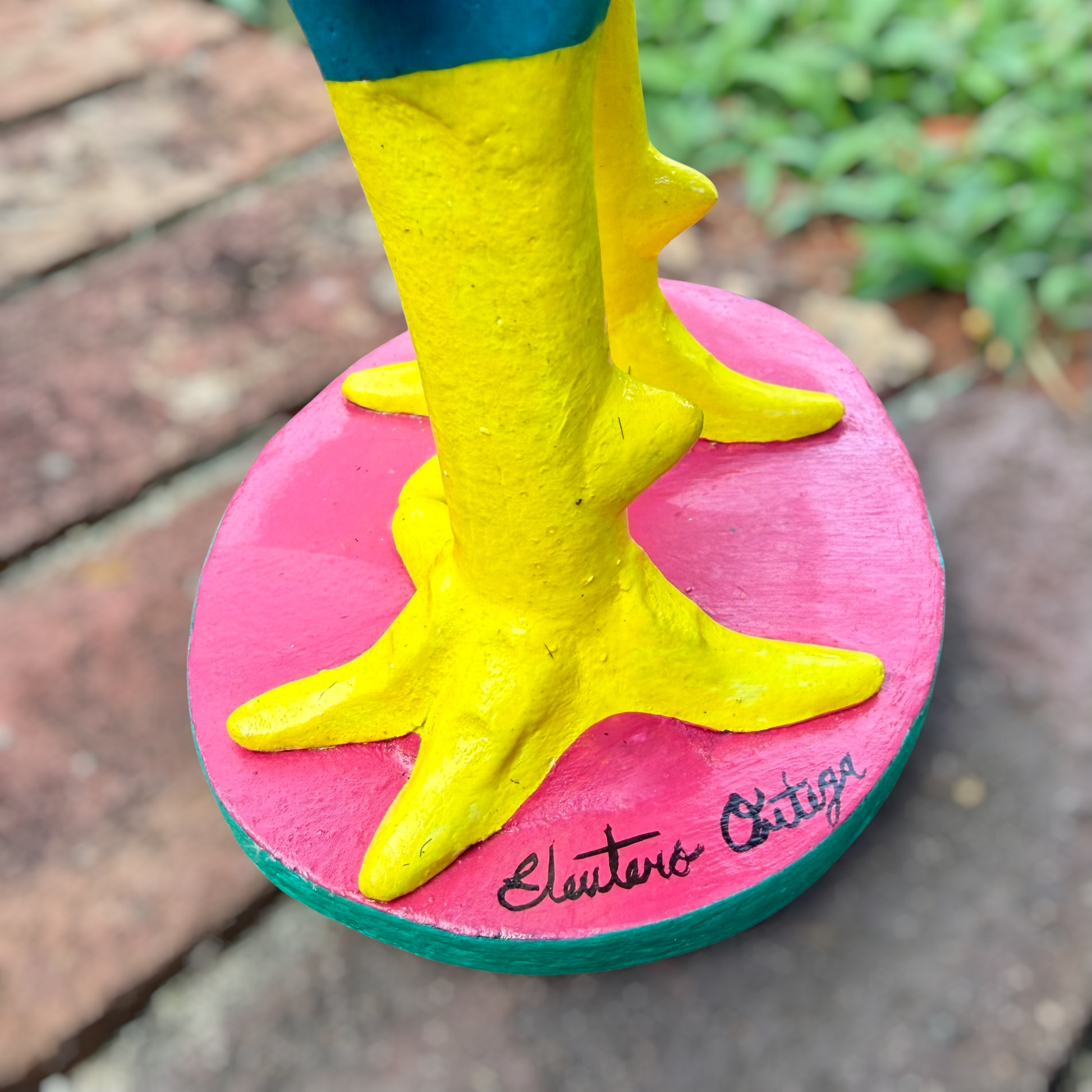 A colorful decorative rooster folk art sculpture with a blue body, yellow legs and beak, and many bright flower ornaments, standing on a pink circular base outdoors on a brick path with a blurred green lawn behind it. Signed by artist in base. 