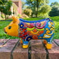 A colorful hand-painted yellow Talavera pig figurine standing on a brick surface with a blurred outdoor garden background, right side view.