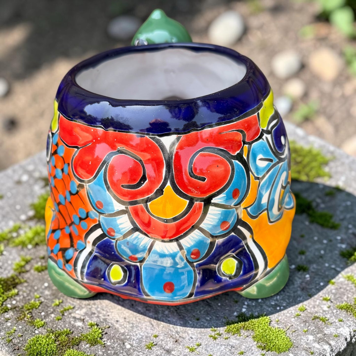 A colorful ceramic turtle-shaped planter with a green head and legs, decorated with vibrant hand-painted patterns in orange, yellow, blue, red, and white, sitting on a mossy stone outdoors. Back of turtle shell.
