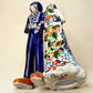 A hand-painted Talavera Day of the Dead bride and groom figurine. The groom is dressed in a blue suit with white patterns, a black bow tie, and an orange-red bow tie accent, wearing a blue hat. Bottom view.