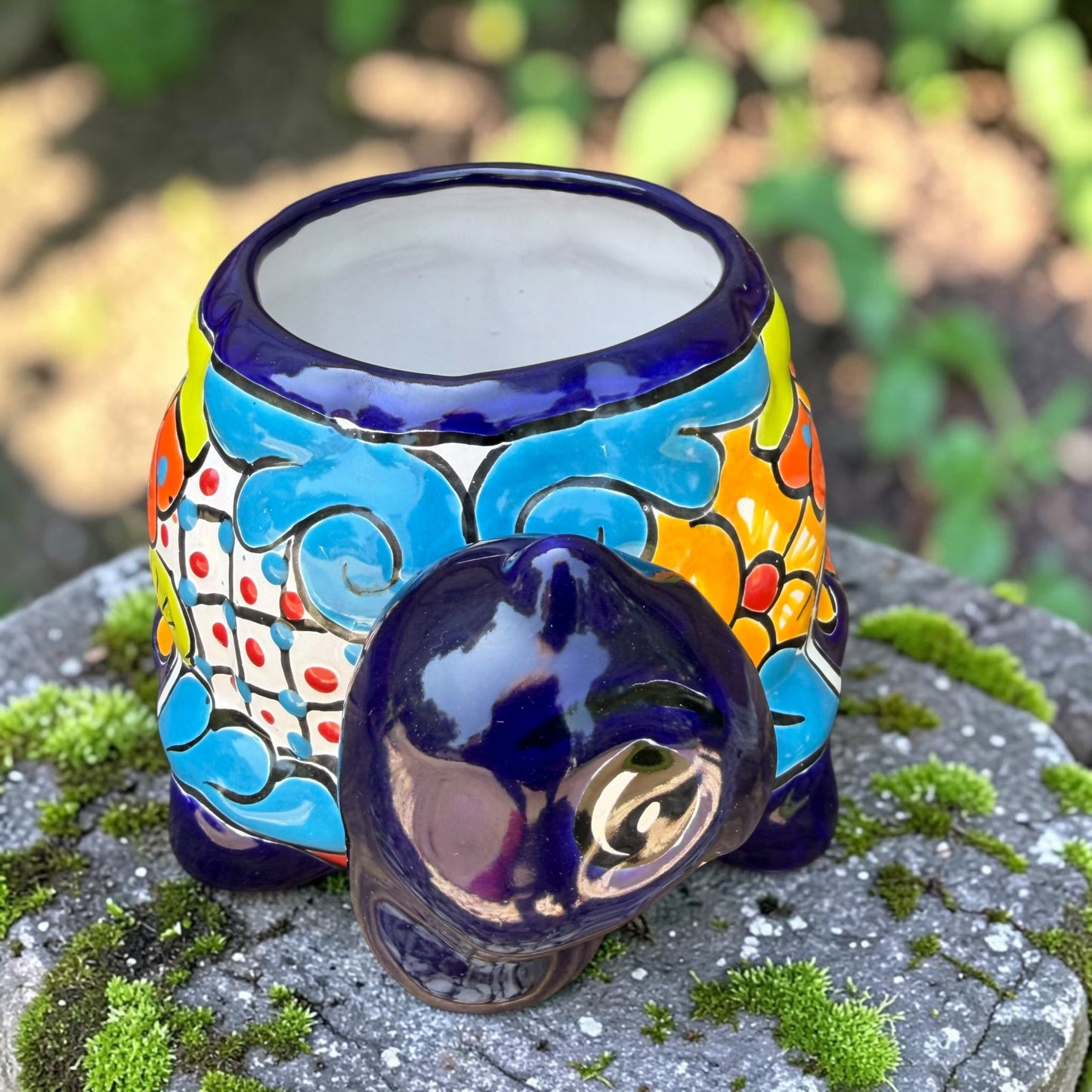 A colorful talavera turtle planter with a hand-painted Mexican-style pattern in orange, blue, yellow, and white, sitting on a mossy stone outdoors. Front of turtle by neck.