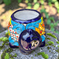 A colorful talavera turtle planter with a hand-painted Mexican-style pattern in orange, blue, yellow, and white, sitting on a mossy stone outdoors. Front of turtle by neck.