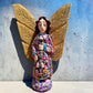 A handmade wood Milagros angel figurine with large gold wings, holding a guitar, dressed in a colorful patterned robe against a textured blue wall.