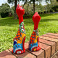 Two colorful hand-painted ceramic cat figurines sitting on a brick ledge outdoors in a grassy park setting. this is their left side view.