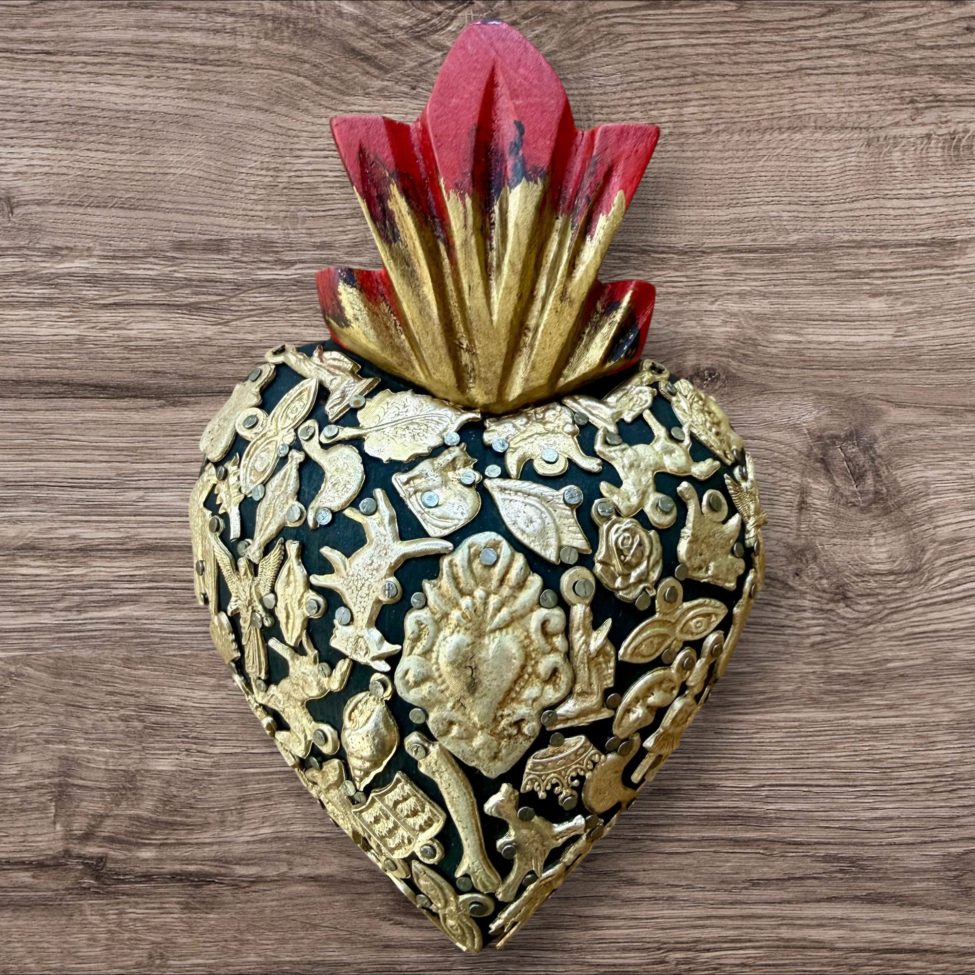 A Mexican Milagros medium heart in black with a gold and red flaming top, covered in antiqued golden charms. 