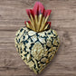 A Mexican Milagros medium heart in black with a gold and red flaming top, covered in antiqued golden charms. 