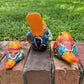 Three hand-painted ceramic bird figurines in yellow, orange  blue, red, and black, displayed on a brick surface with a green grass background back view.