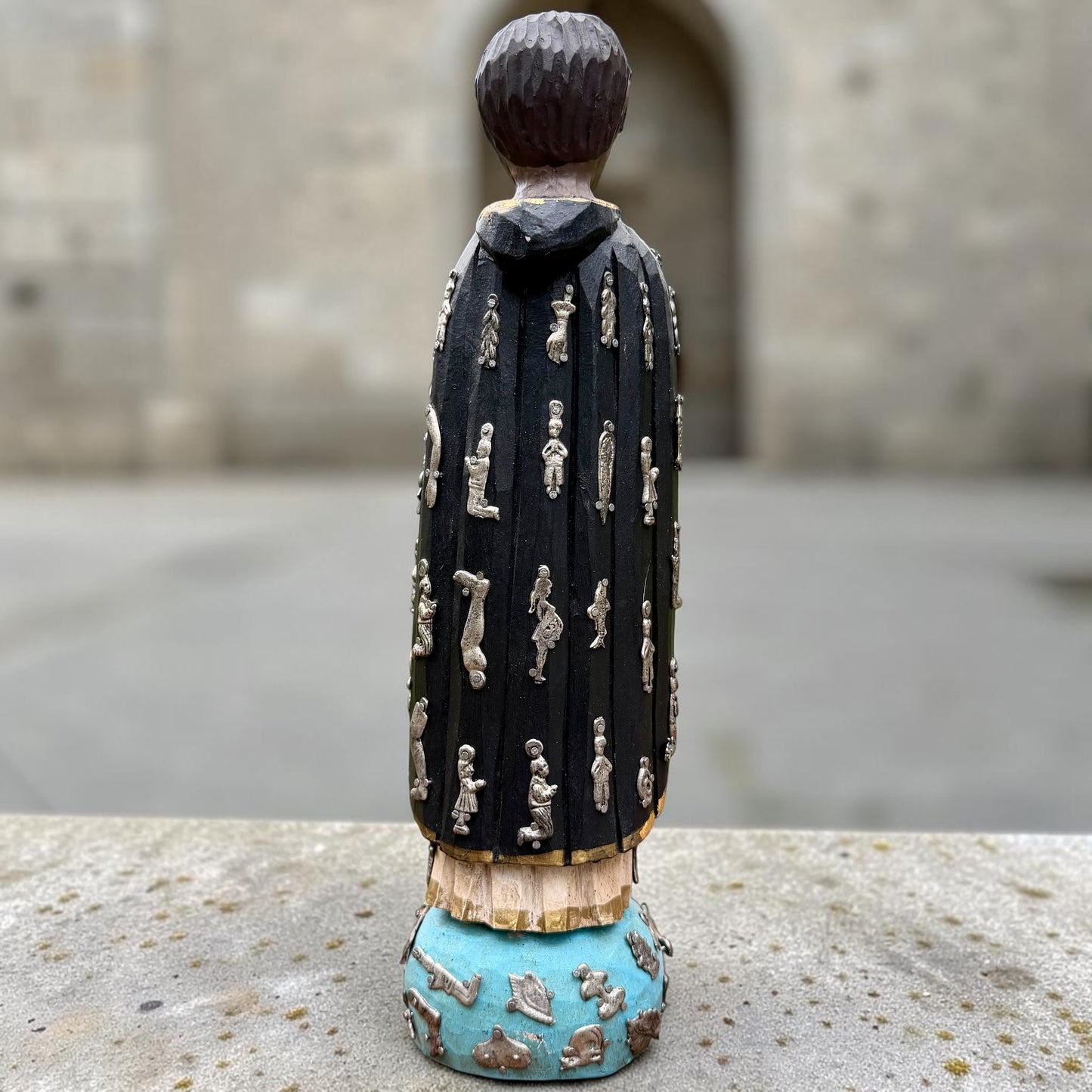 A carved milagros religious figurine of  Saint Martin in ornate black and gold vestments with silver cross details, photographed outdoors in front of a blurred stone archway. Back view of Saint Martin showing milagros charms on robe.