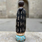 A carved milagros religious figurine of  Saint Martin in ornate black and gold vestments with silver cross details, photographed outdoors in front of a blurred stone archway. Back view of Saint Martin showing milagros charms on robe.